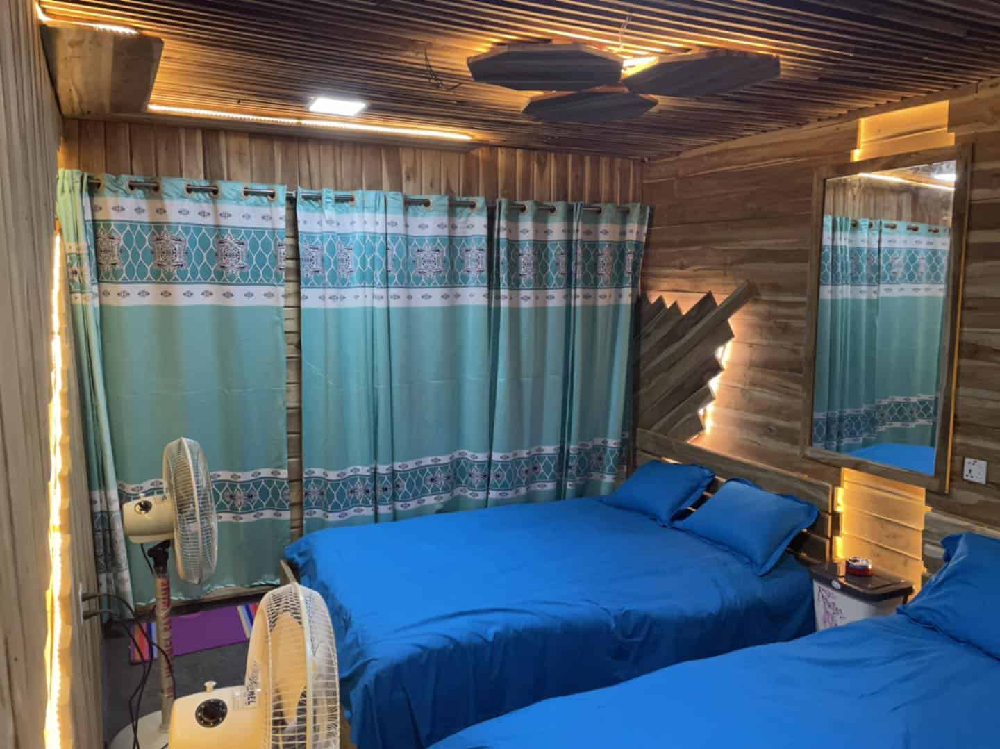 Bright cozy bedroom with wooden walls, blue bedding, patterned curtains, and warm lighting for a relaxing stay.