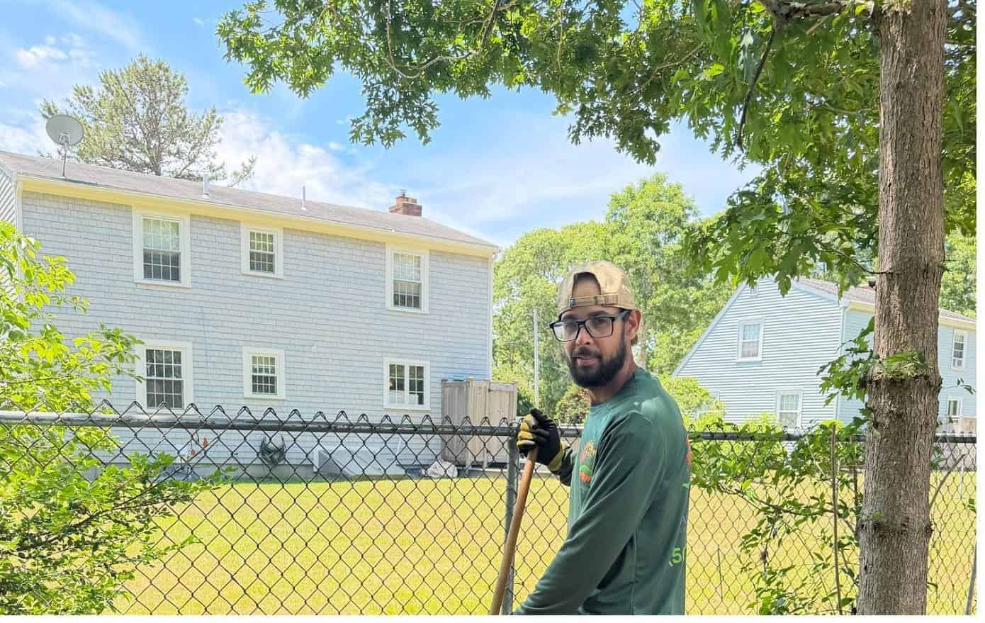Coast Carpentry Home Group crew member in a branded shirt on a remodeling job site in front of a gray cedar shingle Cape Cod colonial
