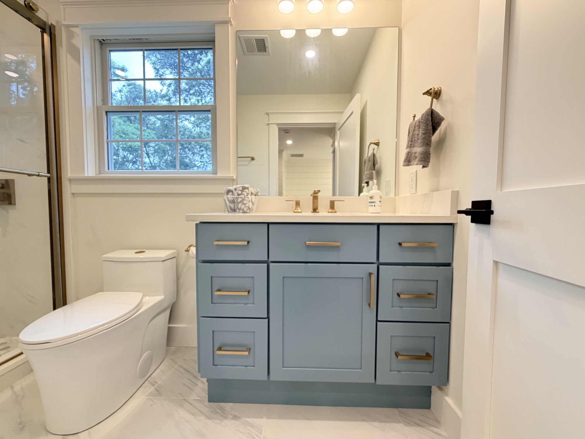 Finished full bathroom built by Coast Carpentry Home Improvement at the 84 Alden Drive new construction on Cape Cod, with a blue vanity, brass hardware, a glass shower and a double hung window on the exterior wall
