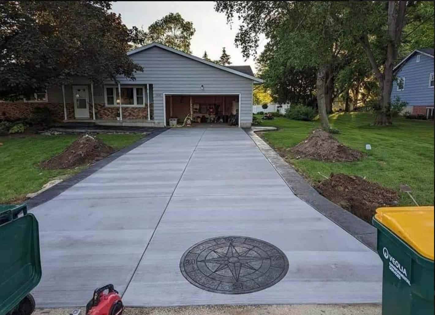 Light‑gray broom‑finish concrete driveway with charcoal border and stamped compass‑rose medallion leading to a single‑car garage.