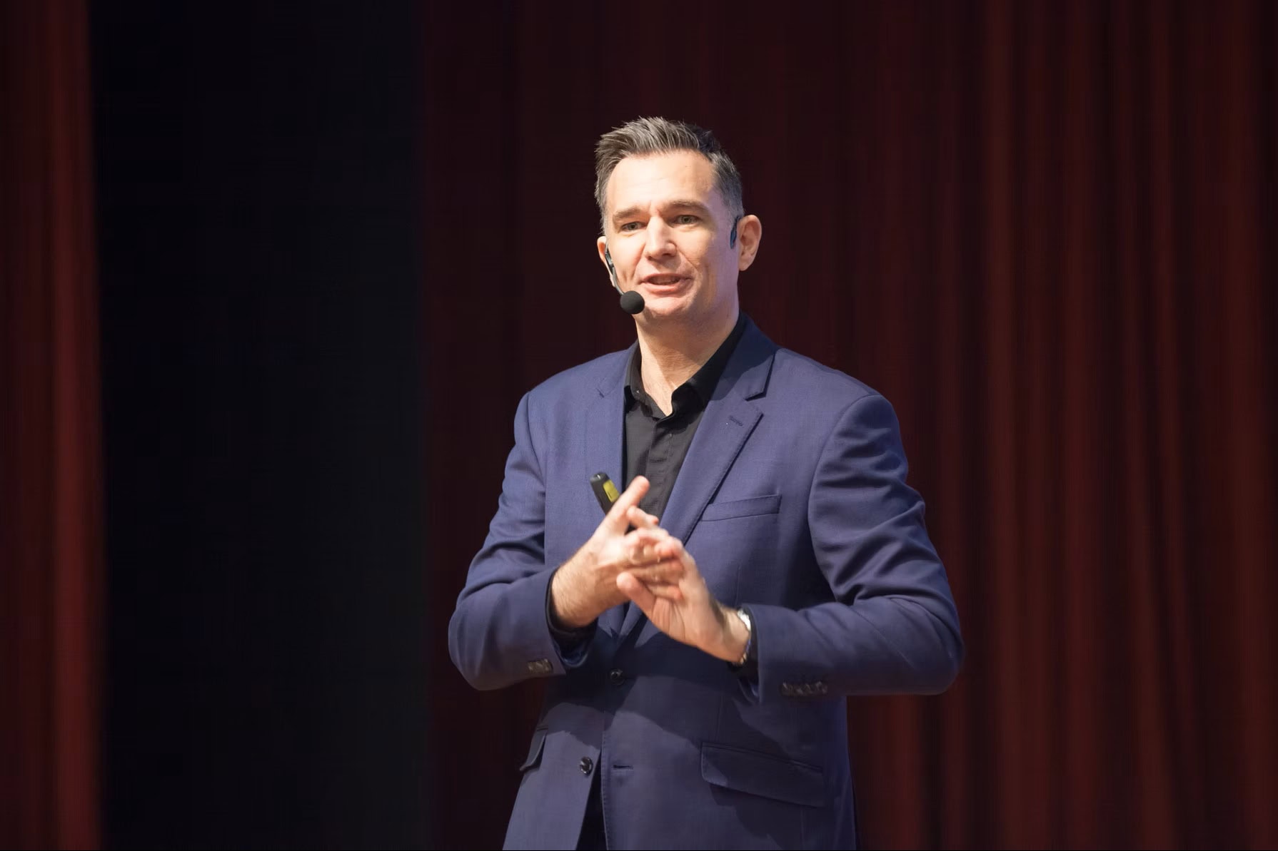 Mark Stuart speaking on stage against a red curtain