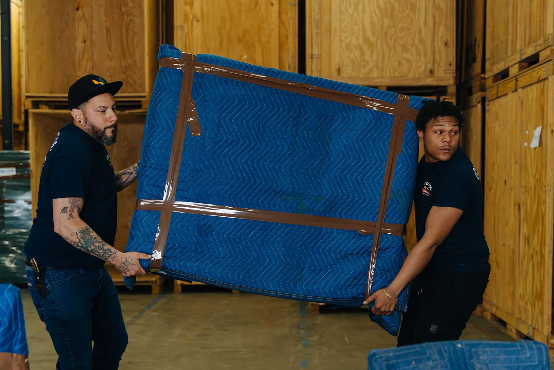 Two movers carefully lift a large blue mattress in a warehouse with wooden crates.