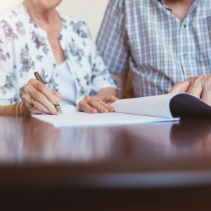An elderly couple, seated at a table, are signing a legal document. Emphasizing the careful consideration required when naming a beneficiary for an IRA.