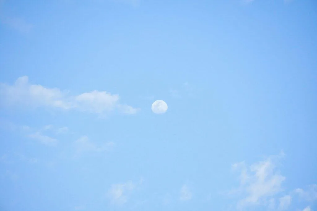 A serene blue sky with the moon visible amidst light white clouds during the day.