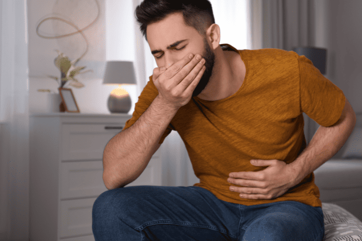 Man seated at home with a hand over his mouth and another on his stomach, feeling nauseous
