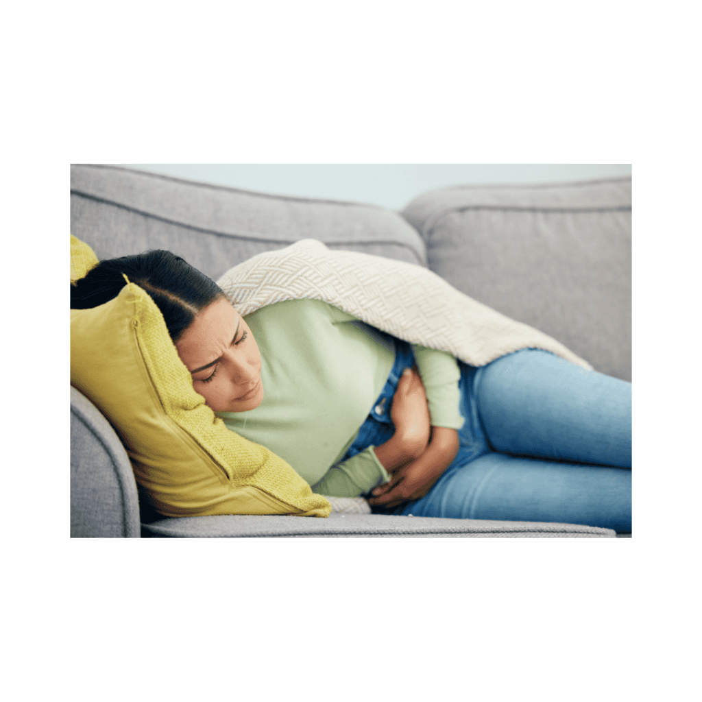 Woman lying on a grey sofa under a blanket, holding her stomach in discomfort from IBS symptoms