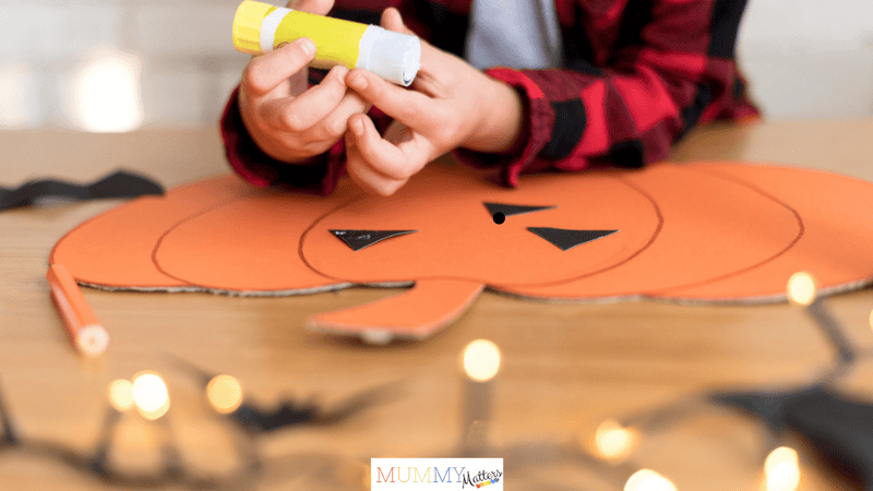 Child making a Halloween pumpkin craft with orange cardboard and a glue stick at home.