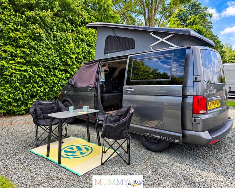 Grey campervan with pop-top roof, outdoor chairs and table set up at a campsite