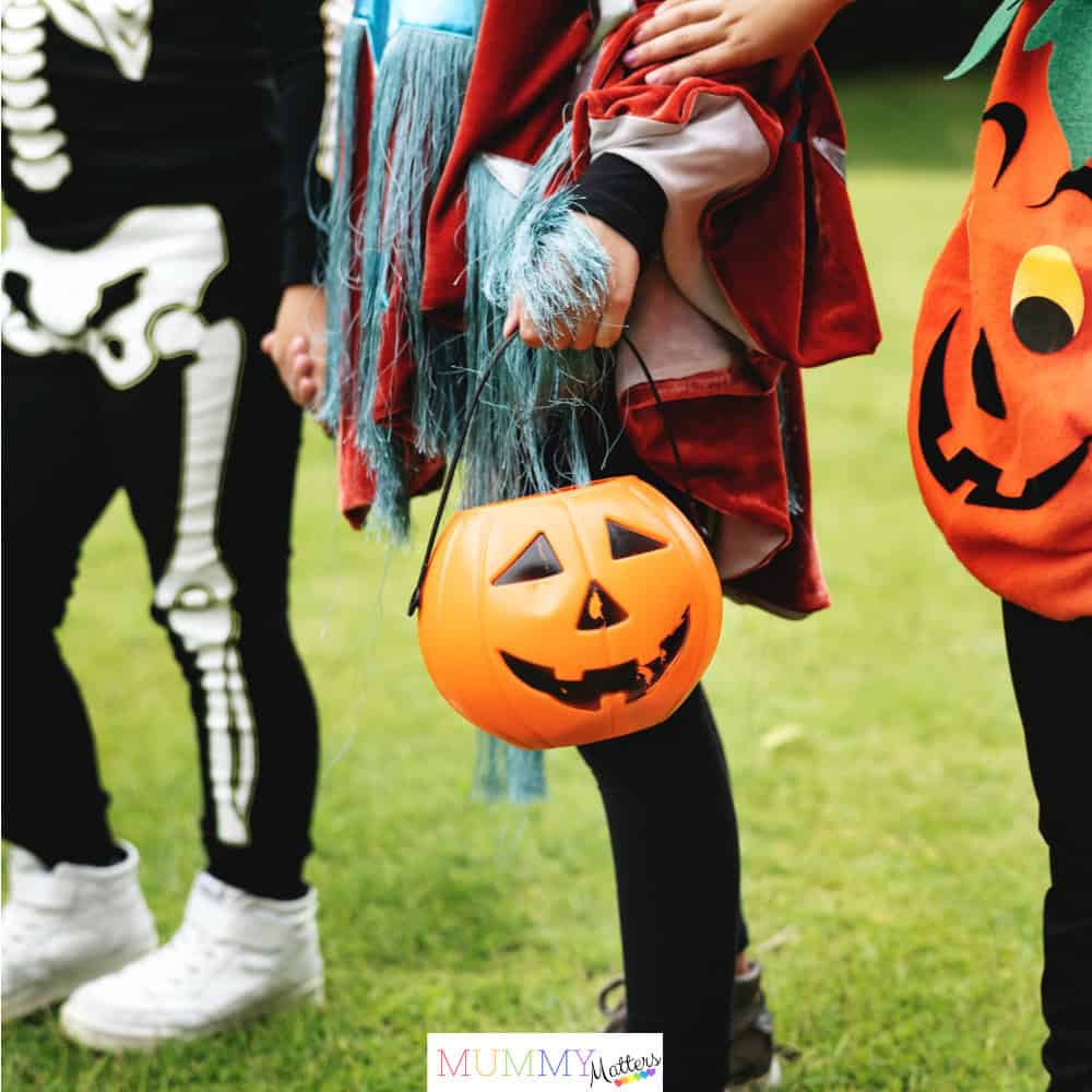Children dressed in colourful Halloween costumes holding hands and carrying a pumpkin trick-or-treat bucket.