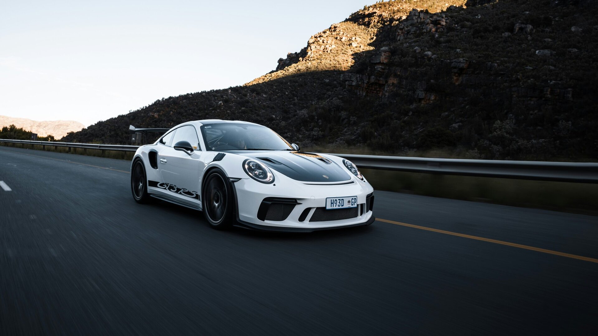 White Porsche sports car driving on a mountain road during sunset, showcasing high performance and sleek design. Perfect for car enthusiasts and luxury vehicle care.