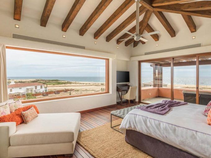 Modern Bedroom With Stunning Ocean Views At Diamante Cabo San Lucas Real Estate Property, Featuring Large Windows, Wooden Ceiling Beams, And Elegant Furnishings.