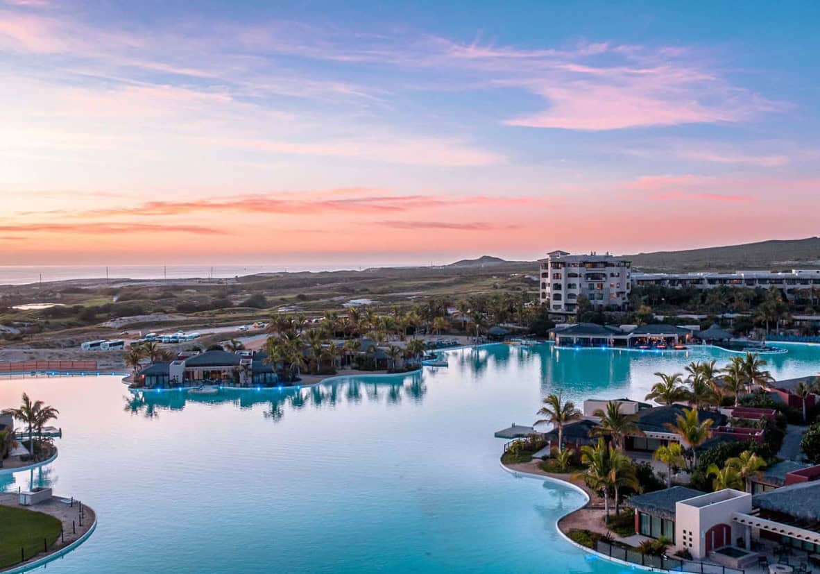 Sunset View Over Diamante Real Estate Showcasing Luxury Residences, Palm Trees, And An Expansive Lagoon-Style Pool In Cabo San Lucas.