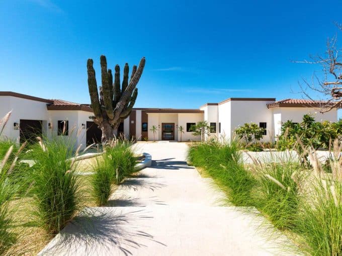 Luxurious Modern Home In Diamante Cabo San Lucas Featuring Desert Landscaping With Cacti And Lush Green Plants, Under A Clear Blue Sky. Diamante Cabo San Lucas Offers Premium Real Estate Options.