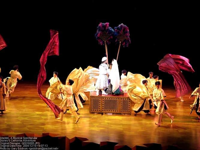 Aladdin and cast dazzle on stage in vibrant costumes, with flowing red banners capturing the magic of Aladdin Pictures live.