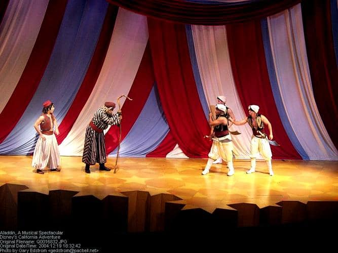 Aladdin and friends light up the stage in vibrant costumes, performing before dazzling red, blue, and white curtains.