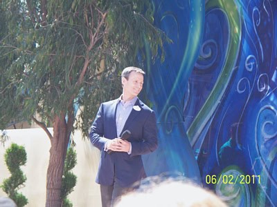 A well-dressed entertainer hosts an outdoor event by a vibrant mural and tree, engaging guests with a microphone on 06/02/2011.