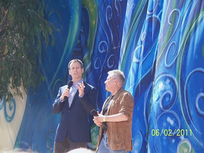 Two performers address the crowd on an outdoor stage, framed by a swirling blue backdrop at a lively theme park event.
