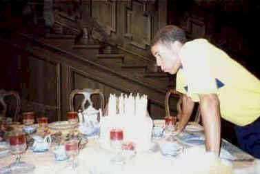 Mickey Mouse in a yellow shirt leans over a birthday cake with lit candles, ready to make a wish backstage at Disneyland.