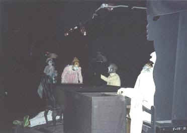 Costumed characters in masks gather around a dimly lit backstage set with a table and chairs, ready for their next performance.