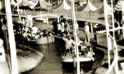 Vintage black-and-white photo of guests boating on a scenic canal at Disneyland, with striped tents shading the ride.