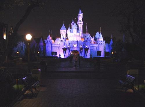Sleeping Beauty Castle aglow at night, viewed from a tree-lined walkway with benches, welcomes Disneyland fans for magical adventures.