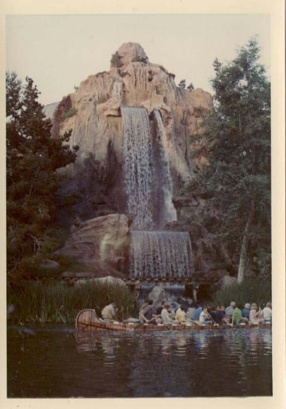 Adventure seekers paddle a canoe past a dramatic double-tiered waterfall at Adventureland, framed by lush, immersive forest.