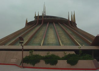Tomorrowland’s iconic cone-shaped building with soaring spires rises above lush greenery beneath a dramatic, cloudy sky.
