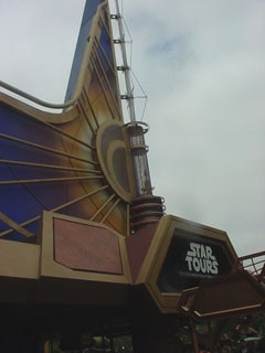 Star Tours entrance in Tomorrowland boasts vibrant, futuristic architecture beneath dramatic cloudy skies, welcoming Disney fans.