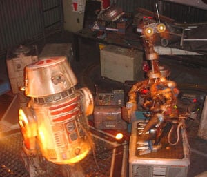 Two robots face off in a Tomorrowland-style setting, surrounded by sci-fi props and themed attraction equipment.