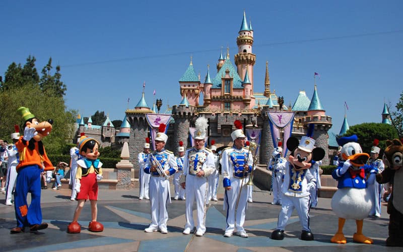 Favorite Disneyland icons and the marching band gather before Sleeping Beauty Castle under sunny skies—theme park magic in action.