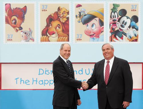 Two executives shake hands before giant USPS stamps spotlighting Mickey Mouse, set against a vibrant Disney-inspired backdrop.