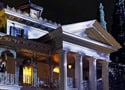 A grand, white-columned mansion aglow at night like Disney’s Haunted Mansion, with eerie yellow-lit windows and shadowy trees behind.