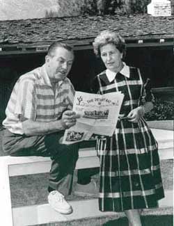 Two guests read a park map together, leaning on a white fence near a themed building in the adventure zone.