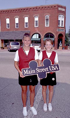 Cast Members in matching costumes hold a "Main Street USA" sign on Main Street with a brick facade in the background.