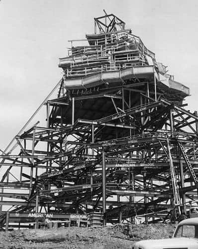 Steel skeleton of a future theme park attraction rises, cranes and beams in action, car parked nearby—exciting thrill ride in progress.