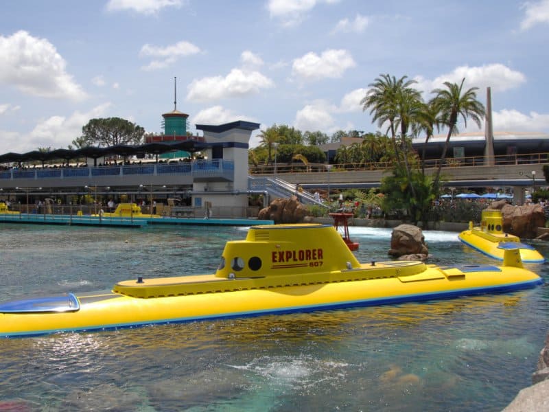 Disneyland's iconic bright yellow "EXPLORER" submarine glides through the lagoon, framed by palm trees and park buildings.