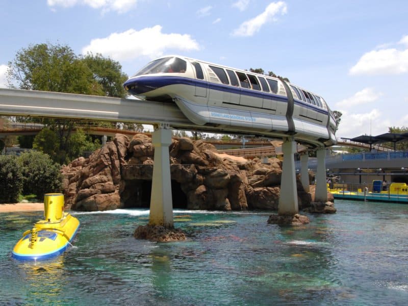 The Disney monorail glides over Epcot’s lagoon above the iconic yellow submarine and themed rock formations on a sunny day.