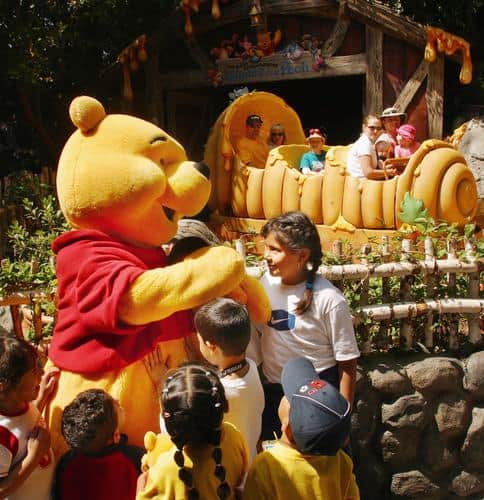 Winnie the Pooh welcomes kids at Disneyland as families enjoy The Many Adventures of Winnie the Pooh ride in the background.