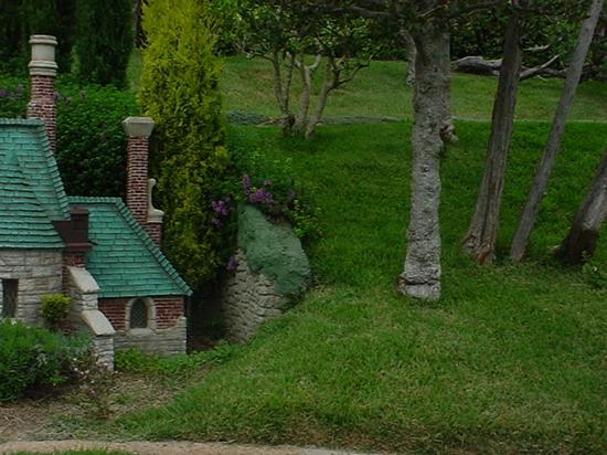 A charming Storybook Land Canal Boats-style cottage with a green roof nestled among tall trees and vibrant grass at Disneyland.