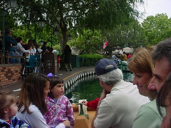 Guests cruise along the Storybook Land Canal Boats at Disneyland, gliding past lush trees and waving onlookers during a photo tour.