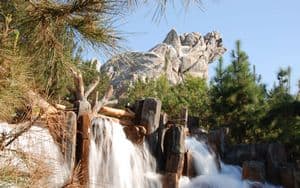 Water rushes over rocks at Disney’s Wilderness Lodge, framed by tall pines and a mountain replica beneath a brilliant sky.