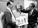 Two Imagineers grin in a Disney workshop, one displaying a character sketch while the other fine-tunes a classic animatronic face.