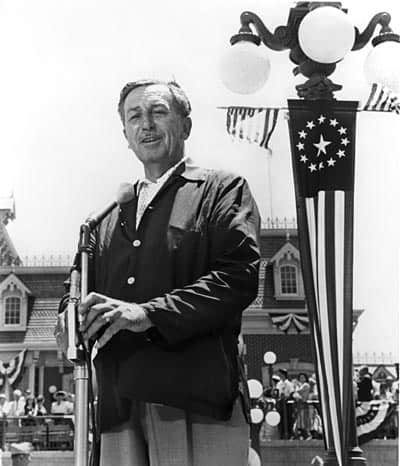 A Disney executive in a suit addresses guests at a podium, with festive decor and a patriotic lamppost evoking Walt Disney’s spirit.