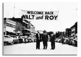 Three men stand by a "Welcome Back Walt and Roy" banner on Main Street, surrounded by crowds and classic Disney vehicles.