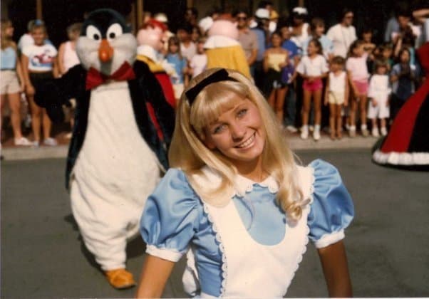 Alice leads a lively parade alongside a playful penguin mascot as excited guests cheer in the theme park background.