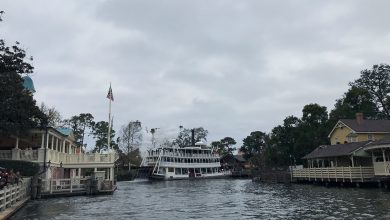 The Mark Twain Riverboat glides past frontier-era wooden buildings under overcast skies at Disneyland.