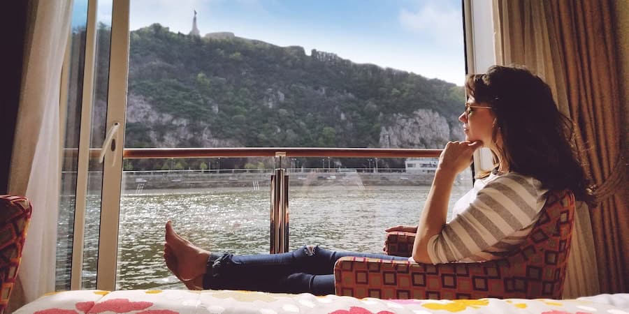 A cruise guest unwinds by a panoramic window, taking in riverside views and a distant hilltop statue during an adventure voyage.