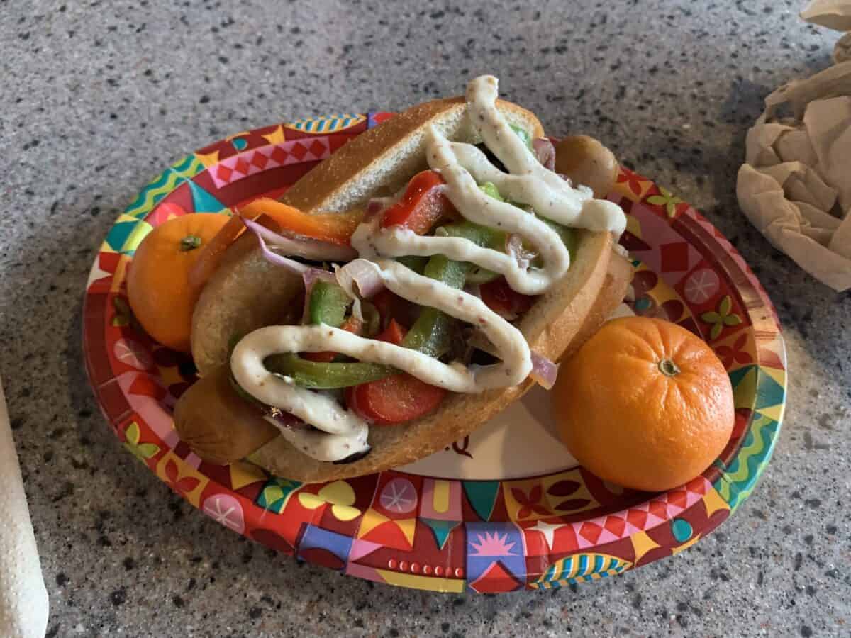 A Disneyland Magic Key-inspired hot dog with veggies and sauce, plus two oranges, on a festive plate—ideal post-park fuel!.