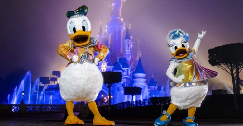Donald Duck and Daisy Duck in vibrant costumes pose together in front of the illuminated Cinderella Castle at night.