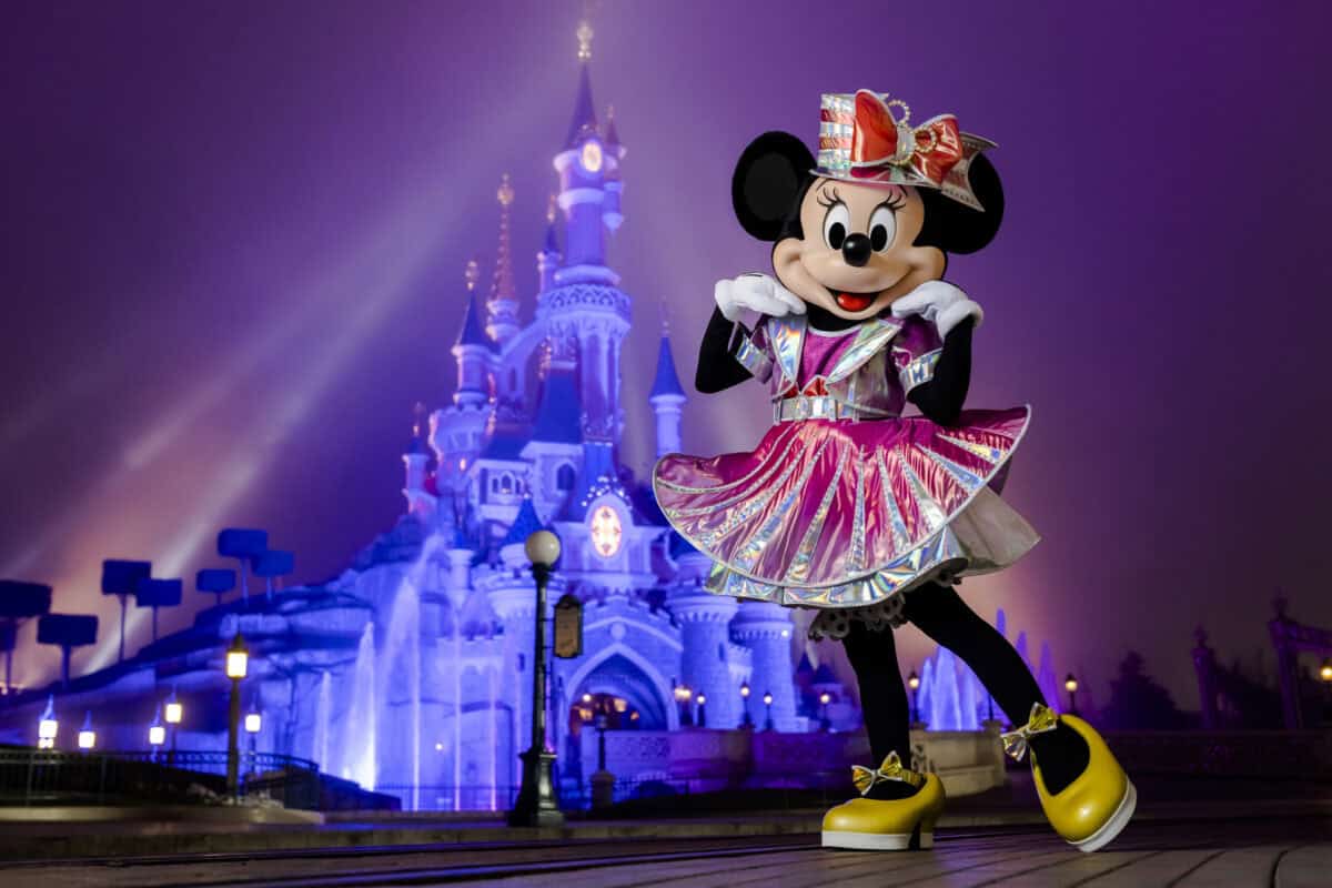 Minnie Mouse sparkles in a shiny dress, posing before the illuminated Disney castle at night—a magical park photo op.